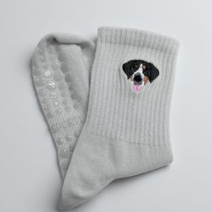 May include: A pair of light gray socks with a dog face embroidered on the cuff. The dog has black, brown, and white markings with a pink tongue. The soles have small, clear, non-slip grips.