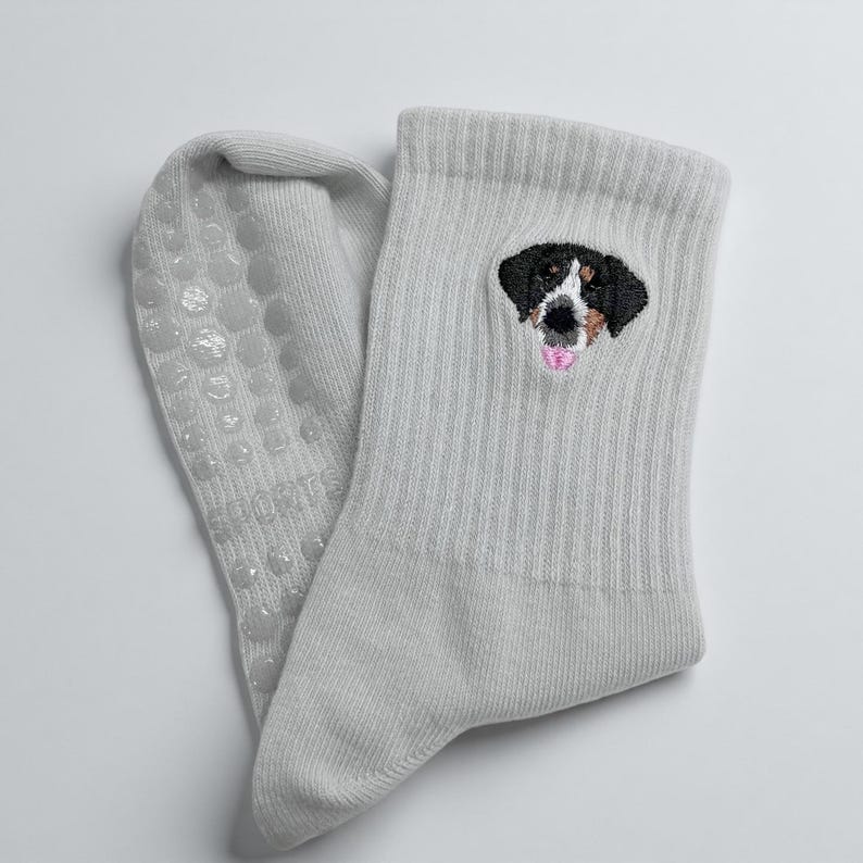 May include: A pair of light gray socks with a dog face embroidered on the cuff. The dog has black, brown, and white markings with a pink tongue. The soles have small, clear, non-slip grips.