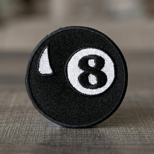 May include: A circular black patch featuring a white number 8 inside a white circle, with a white triangle shape on the left side. The patch is likely for clothing or accessories.
