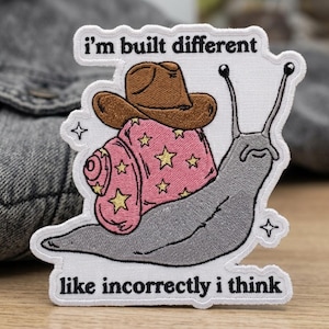 May include: Embroidered patch featuring a cartoon snail wearing a cowboy hat. The snail's shell is pink with gold stars. The text "I'm built different like incorrectly I think" is above and below the snail.