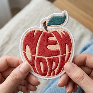 May include: Embroidered patch in the shape of an apple, with the words "NEW YORK" stitched in red. The apple has a white border, a green leaf, and a brown stem. A fun souvenir or accessory.