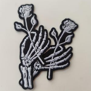 May include: Embroidered patch featuring a skeleton hand holding two roses. The patch has a black background with white and gray detailing. The roses and hand are intricately stitched, creating a detailed and gothic design.