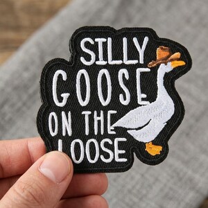 May include: Embroidered patch with the words "SILLY GOOSE ON THE LOOSE" in white on a black background. A cartoon goose wearing a brown hat is featured on the right side of the patch. The patch is held by a hand.