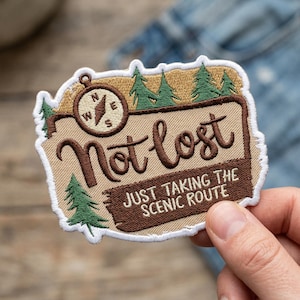 May include: Embroidered patch with the text "Not lost, just taking the scenic route." The patch features a compass, pine trees, and a brown and tan color scheme. The patch has a white border.