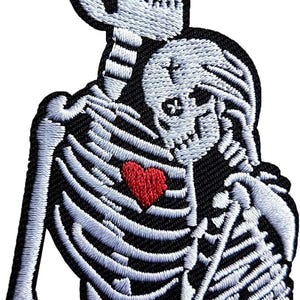 May include: Embroidered patch featuring two skeletons embracing, with a red heart in the center. The skeletons are white with black outlines and are set against a black background. The patch is likely for clothing or accessories.