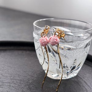 以下が含まれることがあります： ピンク色の結び目のアクセントと、長く細い金色のドロップが付いた、金色のイヤリングのペア。 イヤリングは、暗い背景に対して、水で満たされた透明な模様のガラスの前に展示されています。