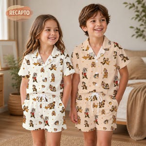 May include: Two children wearing matching pyjama sets. The sets feature a short-sleeved button-up shirt and shorts, both adorned with a cartoon character print. One set is off-white, the other is tan. The pyjamas have a drawstring waist.