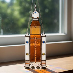 Puede incluir: Un atrapasoles de vidriera con forma de cohete espacial con un diseño detallado. El cohete presenta paneles de vidrio naranja, blanco y azul, suspendidos por una cadena plateada. El atrapasoles se exhibe cerca de una ventana.