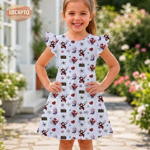 May include: A white dress with a pattern of Spider-Man characters, hearts, and spiderwebs. The dress has short, ruffled sleeves and a fitted bodice. The girl is smiling and standing outside.