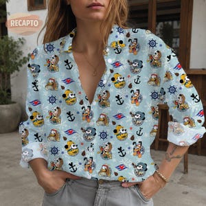 Könnte beinhalten: Ein hellblaues, langärmeliges Hemd mit Knopfleiste und einem Allover-Druck von Disney-Figuren wie Mickey Maus, Donald Duck und Pluto in nautischer Kleidung, durchsetzt mit dunkelblauen Steuerrädern, Ankern und weißen Schneeflocken.