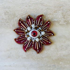 Pode incluir: Broche em forma de flor bordô com um design de olho central. O broche apresenta um olho vermelho e branco, rodeado por contas brancas e douradas e detalhes dourados. As pétalas são contornadas em dourado.