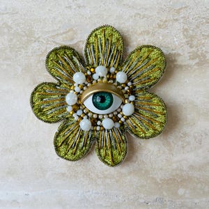 Puede incluir: Broche con forma de flor y diseño de ojo. Los pétalos están bordados en verde y dorado, con un ribete metálico dorado. El ojo presenta un iris verde, rodeado de cuentas blancas y doradas. Un accesorio artístico y distintivo.