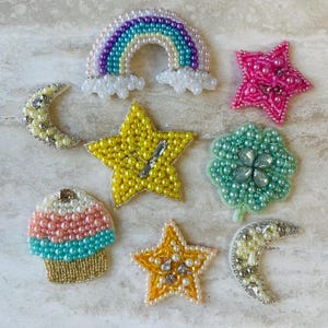 May include: A collection of beaded embellishments in various shapes, including a rainbow, stars, a crescent moon, a cupcake, and a flower. The embellishments feature a variety of colors, including pink, yellow, blue, and white, and are likely used for crafting or decorating.