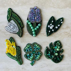 Könnte beinhalten: Eine Sammlung von sechs handgefertigten, bestickten Broschen in verschiedenen Blumen- und Blattdesigns. Die Broschen weisen aufwendige Perlenarbeiten in Grün-, Weiß-, Gelb- und Lilatönen mit Perlenakzenten auf. Die Designs umfassen einen Schmetterling, eine Blume, Blätter und ein vierblättriges Kleeblatt.