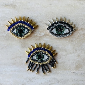 Peut inclure: Trois écussons en forme d'œil perlés. Chaque œil présente un iris détaillé avec des perles blanches, vertes et noires. Les yeux sont entourés de pointes dorées ou argentées et de sourcils perlés bleus.