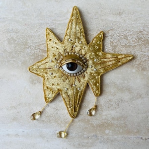 Peut inclure: Décoration dorée en forme d'étoile avec un œil central. L'œil est brun avec des détails dorés, entouré de petites perles blanches. L'étoile a une surface texturée et trois ornements en forme de goutte suspendus.