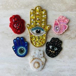 Könnte beinhalten: Eine Sammlung von Hamsa-Hand-Anstecknadeln aus Perlen in verschiedenen Farben. Die Anstecknadeln umfassen Rot, Blau, Rosa, Schwarz, Weiß und Gold. Die zentrale goldene Anstecknadel weist ein böses Augendesign auf. Jede Anstecknadel ist aufwendig mit Perlen und Pailletten verziert.