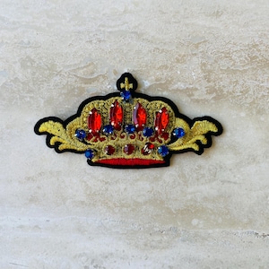 May include: Embroidered crown patch with gold thread, black backing, and red and blue jewel accents. The crown features a detailed design with a regal aesthetic, suitable for embellishing clothing or accessories.