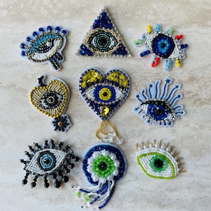 Może przedstawiać: Kolekcja naszywek w kształcie oka z koralików w różnych wzorach. Naszywki charakteryzują się skomplikowanymi koralikami w kolorze niebieskim, złotym, białym i zielonym, niektóre w kształcie serc i trójkątów. Te dekoracyjne naszywki są idealne do dodania niepowtarzalnego charakteru odzieży lub akcesoriom.