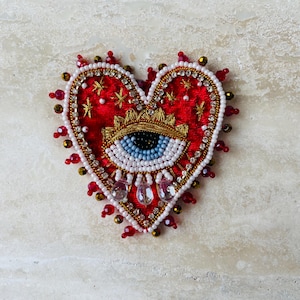 May include: A heart-shaped embroidered brooch with a red velvet base. The design features a gold crown, an eye with blue iris, and star accents. The brooch is embellished with white, red, and gold beads, and clear crystal teardrops.