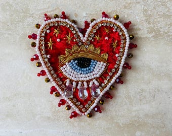 Op de afbeelding: Een hartvormige, geborduurde broche met een rode fluwelen basis. Het ontwerp heeft een gouden kroon, een oog met blauwe iris en steraccenten. De broche is versierd met witte, rode en gouden kralen en heldere kristallen druppels.