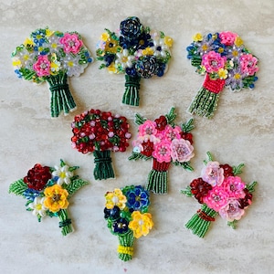 Könnte beinhalten: Eine Sammlung von Perlenblumensträußen in verschiedenen Farben, darunter Rosa, Rot, Gelb, Blau und Weiß. Jeder Strauß ist sorgfältig mit Perlen und Pailletten gefertigt und mit grünen Stielen gebunden. Die Sträuße sind auf einer hellen Oberfläche angeordnet.