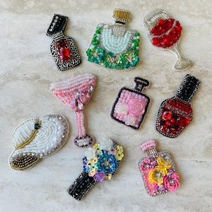 Może przedstawiać: Kolekcja broszek z koralików w różnych kształtach, w tym kieliszek do wina, butelki perfum, kieliszek do koktajli, kapelusz i bukiet. Broszki charakteryzują się różnorodnością kolorów, w tym czerwonym, różowym, zielonym i białym, z misternymi koralikami i zdobieniami.