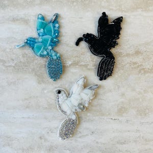 May include: Three beaded hummingbird ornaments in blue, black, and white. The birds have detailed wings and tails, with bead and sequin embellishments. These ornaments are likely for decorative purposes.