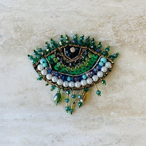 May include: An eye-shaped brooch with a black base, embellished with green sequins, beads, and faux pearls. The brooch features a central crystal and dangling green bead accents. The design incorporates shades of green, blue, and white, with gold-toned accents.