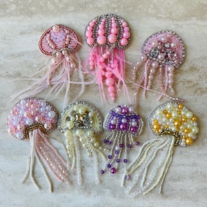 Puede incluir: Colección de broches de medusas hechos a mano. Cada broche presenta una cúpula de cuentas en tonos rosas, morados y amarillos, con tentáculos de cuentas colgantes y detalles de plumas. Los broches están adornados con perlas y cristales.