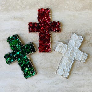 May include: Three decorative crosses in red, green, and white. The red cross is covered in sequins and outlined with rhinestones. The green cross is also sequined and rhinestone-trimmed. The white cross features floral embellishments and pearl accents.
