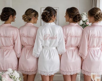 Puede incluir: Cinco batas de satén con ribete de encaje en las mangas y el dobladillo. Cuatro batas son de color rosa claro con la palabra Bridesmaid bordada en la espalda en cursiva. La bata central es blanca con la palabra Bride bordada en cursiva.