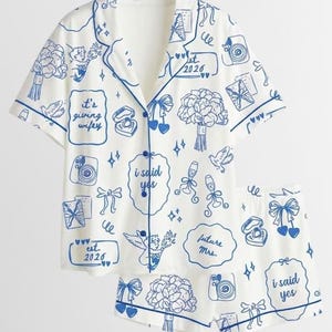 May include: A white short-sleeve button-down pajama shirt and matching shorts set featuring a blue line-art print of wedding-themed icons, including bouquets, rings, cameras, and text reading "it's giving wifey," "i said yes," and "future mrs."