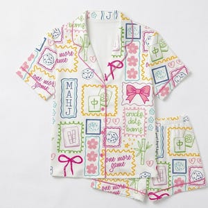 Op de afbeelding: Een witte pyjamaset bestaande uit een shirt met korte mouwen en knoopsluiting en een bijpassende short. De stof is bedrukt met een kleurrijk patroon van mahjongtegels, dobbelstenen, roze kersenbloesems, strikjes, bamboe, hartjes en de tekst "one more game".