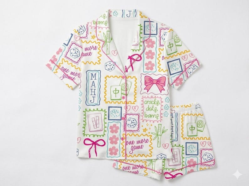 May include: A white pajama set featuring a short-sleeved button-down shirt and matching shorts. The fabric is covered in a colorful pattern of mahjong tiles, dice, pink cherry blossoms, bows, bamboo stalks, hearts, and the text "one more game".