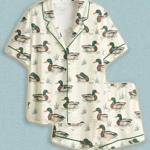 Könnte beinhalten: Cremefarbenes Pyjama-Set mit einem sich wiederholenden Muster von Enten auf dem Wasser. Das kurzärmelige Oberteil und die Shorts sind mit grünen Paspeln versehen. Die Enten sind in Braun-, Grün- und Weißtönen dargestellt.
