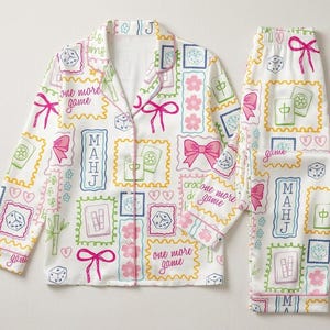 May include: A white pajama set featuring a long-sleeved button-down shirt and matching pants. The fabric is covered in a colorful print of mahjong tiles, dice, pink bows, cherry blossoms, and the text "one more game" and "MAHJ" in frames.
