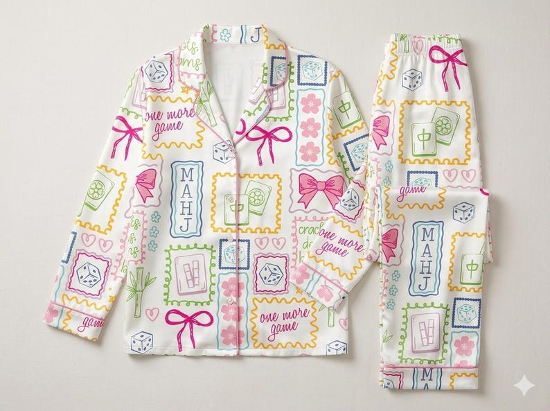 May include: A white pajama set featuring a long-sleeved button-down shirt and matching pants. The fabric is covered in a colorful print of mahjong tiles, dice, pink bows, cherry blossoms, and the text "one more game" and "MAHJ" in frames.