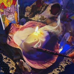 May include: A lit candle in a seashell, with a tarot card and a skull in the background. The candle is white and has a flame. Incense is burning next to the candle. The tarot card shows an angel with a yellow background.