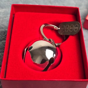 Puede incluir: Un llavero de cascabel de metal plateado con una campana redonda y una pequeña etiqueta ovalada grabada con el texto 2000 RING IN THE MILLENNIUM. El llavero se presenta dentro de una caja cuadrada forrada con tela de terciopelo rojo brillante.
