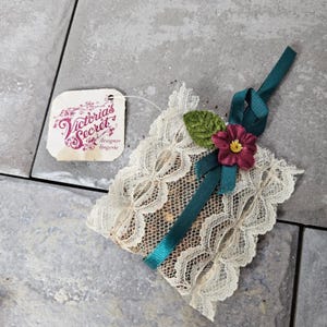 May include: A square sachet covered in cream-colored lace with a dark teal ribbon tied in a bow. A small dark pink flower and a green fabric leaf are attached to the ribbon. A white tag attached by a string reads Victoria's Secret designer lingerie.