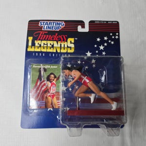 May include: A Starting Lineup Timeless Legends 1996 Edition action figure of Florence Griffith Joyner in a red and white track uniform. The packaging features the American flag and the text "Florence Griffith Joyner".