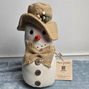 May include: A fabric snowman figurine wearing a tan burlap hat adorned with small decorative berries and pinecones. The snowman features a bright orange nose, black button eyes, a burlap bow tie, and three dark brown buttons on its torso.