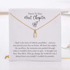 May include: A gold chain necklace with an oval opal pendant displayed on a white card featuring a rocket icon and the text: Here's To Your Next Chapter. The card includes a message about opal being the stone of infinite possibility.