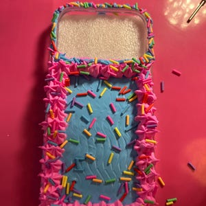 Può includere: Una custodia trasparente per telefono decorata con glassa rosa, glassa blu e confettini colorati. La custodia ha una forma rettangolare con angoli arrotondati e un centro trasparente.