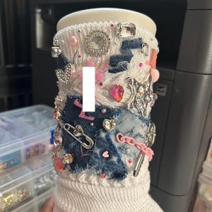 May include: A white boot with denim patches, embellished with rhinestones, pearls, and silver charms. The boot features a safety pin, a pink chain, and various decorative elements. The design incorporates a mix of textures and colors.