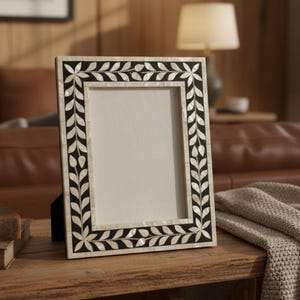 Op de afbeelding: Een rechthoekige fotolijst met een zwart-witte bladeren design rand. De lijst is gemaakt van een lichtgekleurd materiaal en heeft een zwarte achterkant. De lijst wordt op een houten tafel getoond.