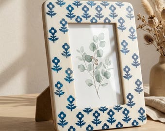 Op de afbeelding: Een rechthoekige fotolijst met een crèmekleurige rand versierd met blauwe bloempatronen. De lijst bevat een aquarelprint van eucalyptusbladeren. De lijst wordt ondersteund door een houten standaard.