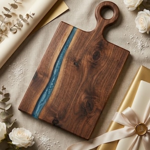 Puede incluir: Una tabla de cortar rectangular de madera con un asa. La tabla es de madera marrón oscuro con un río de resina azul en el centro. El asa tiene una abertura circular. La tabla está rodeada de rosas blancas y un regalo envuelto.