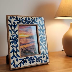 Pode incluir: Moldura de madeira com um design floral azul e branco. Contém uma foto de uma cena de praia ao pôr do sol. A moldura está sobre uma superfície de madeira.